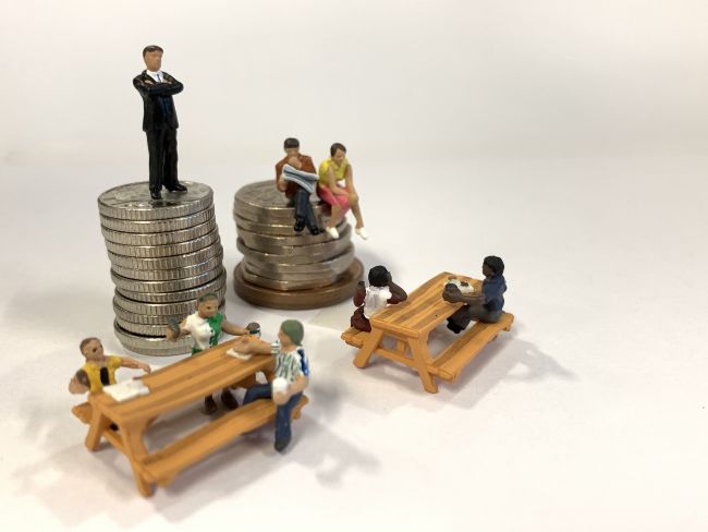 small model figures sitting on benches beside a stack of coins